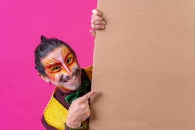 Clown with white facial makeup showing a sign on a pink background, pointing