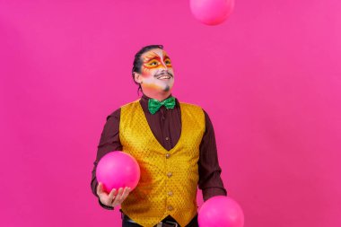 Clown with white facial makeup on a pink background, smiling juggling balls