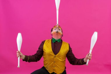 Clown with white facial makeup on a pink background, smiling juggling clubs on his face