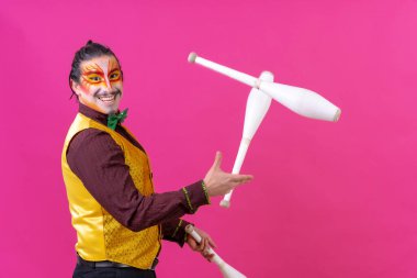 Clown with white facial makeup on a pink background, smiling juggling clubs and looking at camera