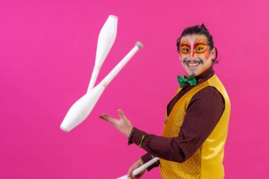 Clown with white facial makeup on a pink background, smiling juggling clubs and looking at camera