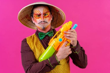 Clown with white facial makeup on a pink background, playing with toy guns