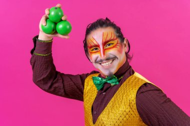 Clown with white facial makeup on a pink background, juggling green balls
