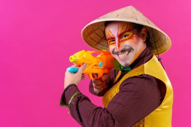 Clown with white facial makeup on a pink background, smiling with toy guns