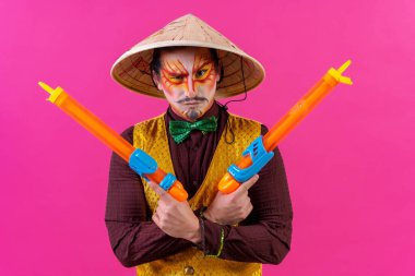 Clown with white facial makeup on a pink background, with toy guns
