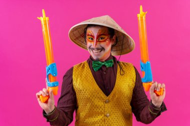 Clown with white facial makeup on a pink background, smiling with toy guns