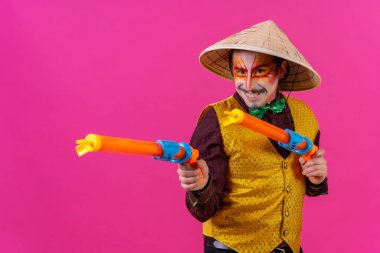 Clown with white facial makeup on a pink background, with toy guns and a Chinese hat