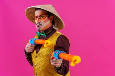 Clown with white facial makeup on a pink background, with toy guns and a Chinese hat