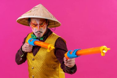 Clown with white facial makeup on a pink background, with toy guns and a Chinese hat