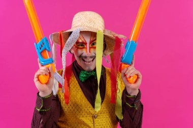 Clown with white facial makeup on a pink background, with toy guns and a hat