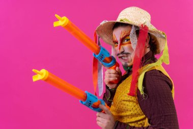 Juggler with white facial makeup on a pink background, smiling with toy guns and hat
