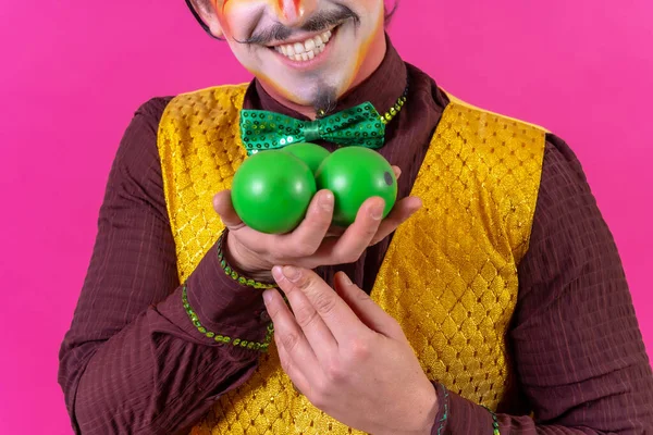 Portrait of clown with white facial makeup on a pink background, juggling green balls