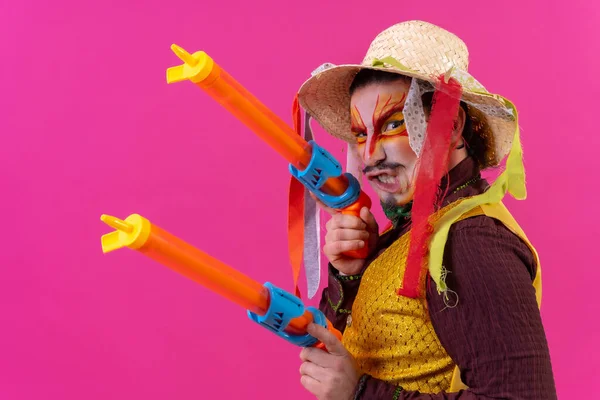 Juggler with white facial makeup on a pink background, smiling with toy guns and hat