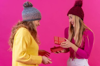 İki genç sarışın beyaz kadın gülümsüyor pembe bir arka planda izole edilmiş arkadaşının hediyesine bakıyorlar.