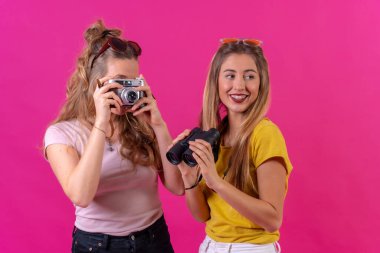 Pembe arka planda izole edilmiş kamera ve dürbünlü iki sarışın beyaz turist portresi.