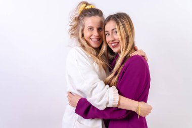 Beyaz bir arka planda sarılıp gülümseyen iki sarışın kadın arkadaş.