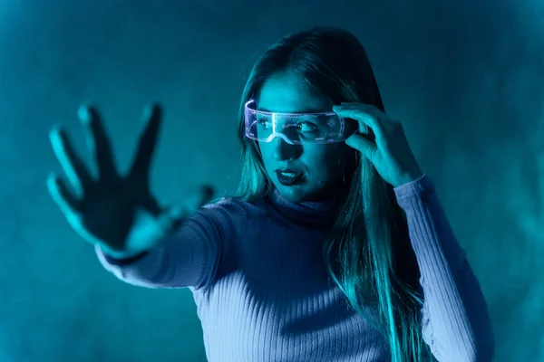 Blonde woman with futuristic glasses gesturing against a blue background, making the stop gesture