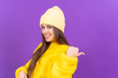 Attractive woman smiling pointing fingers at copy space on purple background, wool sweater