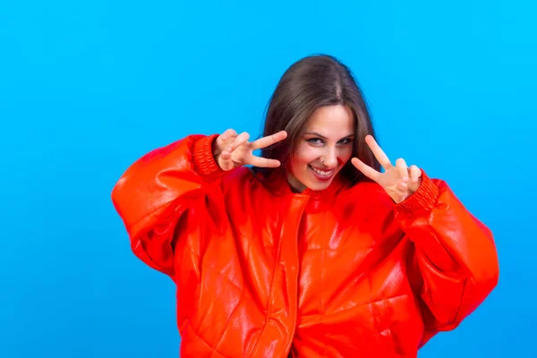 Young woman over blue wall smiling and showing victory sign with red windbreaker