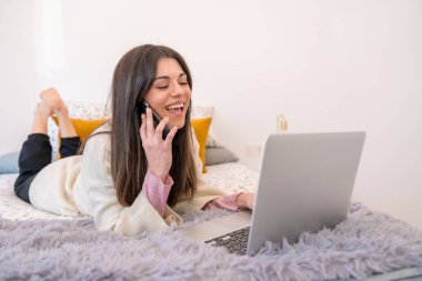 Yatakta dizüstü bilgisayarla çalışan, evden işe giden, tele çalışan, telefonda gülümseyen bir kadın.