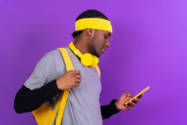 Mor arka planda sırt çantası ve sarı kulaklıklı siyah etnik adam, öğrenci konsepti.