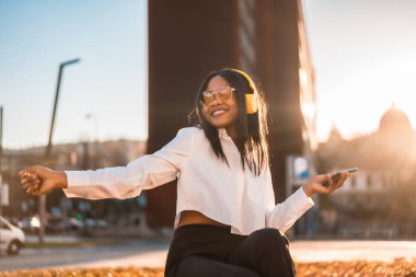 Şehirde günbatımında kulaklıkla müzik dinleyen Afrikalı Amerikalı bir kadın. İnternetten müzik ve dans.