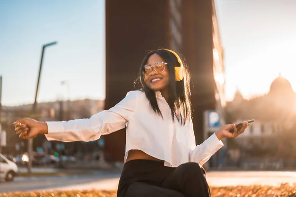 African american woman listening to music with headphones at sunset in the city, music online and dancing