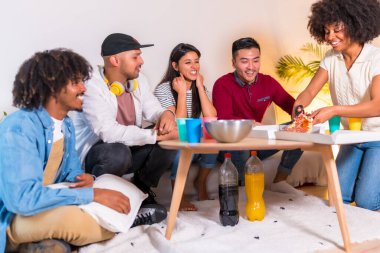 Bir grup çok ırklı arkadaş bir kanepede pizza yiyor ve ev partisinde meşrubat içiyor, kız pizzayı kesiyor ve dağıtıyor.