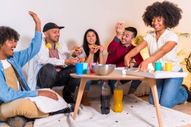 Bir grup çok ırklı arkadaş bir kanepede pizza yiyor ve bir ev partisinde meşrubat içiyor, eğleniyor ve gülümsüyor.