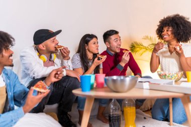 Bir grup çok ırklı arkadaş bir kanepede pizza yiyip gazoz içiyorlar, pizza yiyorlar ve şakalar yapıyorlar.