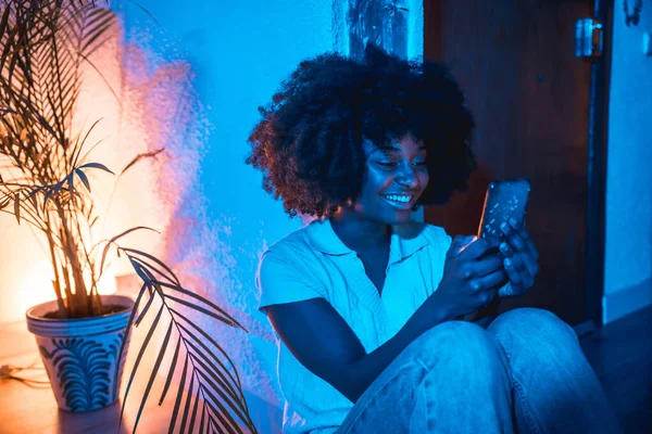 Kobieta Afro Włosy Siedzi Domu Podłodze Patrząc Telefon Nocy Niebieskim — Zdjęcie stockowe