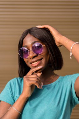 Şık Yaz Turisti: Güneş Gözlüklü Bir Siyah Kadın Portresi ve Kahverengi Duvara Karşı Yeşil Tee Poz