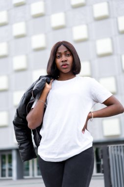 Şehir modası: Genç siyahi bir kadın modern gri bir bina olarak şık, kentsel bir zemin olarak, şık bir beyaz tişört ve siyah pantolon içinde poz veriyor