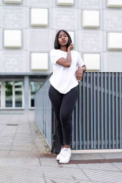 stock image Chic and stylish: A portrait of a young black woman in a white tee and black pants against a grey building