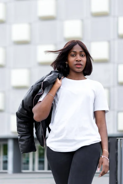 Fashionable city vibes: A young black woman strikes a pose in a stylish white tee and black pants, with a grey building as a chic urban backdrop