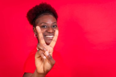 Genç Afrikalı Amerikalı kadın kırmızı bir arka planda izole edilmiş zafer jesti ile gülümsüyor, stüdyo çekimi