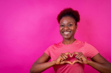 Genç Afrikalı Amerikalı kadın pembe bir arka planda izole edilmiş gülümsüyor ve kalp hareketi, stüdyo çekimi