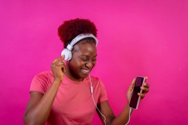 Genç Afrikalı Amerikalı bir kadın pembe arka planda izole edilmiş, kulaklık takıyor, dans ediyor, stüdyo çekimi yapıyor.