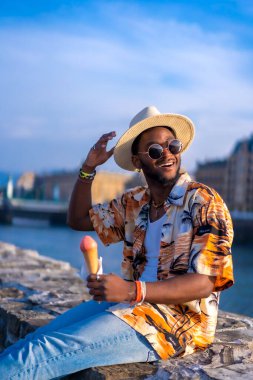 Siyahi bir etnik adamın portresi sahilde yaz tatilinin tadını çıkarıyor dondurma yiyor, nehir kenarında oturuyor.