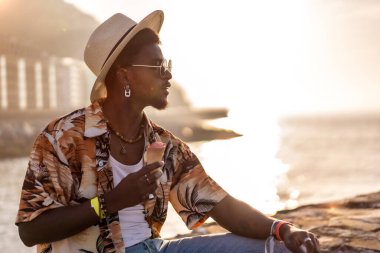 Siyahi bir etnik adamın günbatımı plajda dondurma yiyerek yaz tatilinin tadını çıkarıyor.