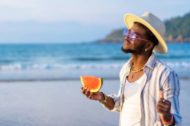 Siyahi bir etnik adamın portresi plajda karpuz yiyip özgürlüğün tadını çıkarıyor.