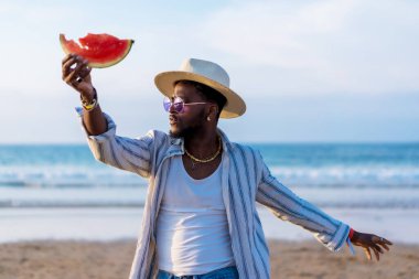 Siyahi etnik adam yaz tatilinin tadını deniz kenarında karpuz yiyerek çıkarıyor.