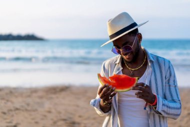 Siyahi bir etnik adamın portresi deniz kenarında karpuz yiyerek yaz tatilinin tadını çıkarıyor.