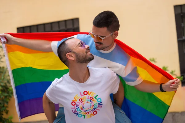 同性恋男性情侣骑在背上亲吻对方在骄傲的聚会上与彩虹旗 Lgbt概念 — 图库照片
