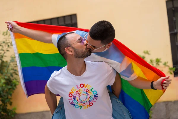 同性恋男性情侣骑在背上亲吻对方在骄傲的聚会上与彩虹旗 Lgbt概念 — 图库照片