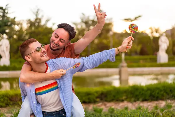 在城市的日落时分 同性恋男友在公园里玩耍 其中一个爬到后面 一边吃棒棒糖一边跑 Lgbt概念 — 图库照片