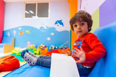 Mavi bir kanepede oturan bir çocuk masal kitabı okuyor. Anaokulunun içinde kameraya bakıyor.