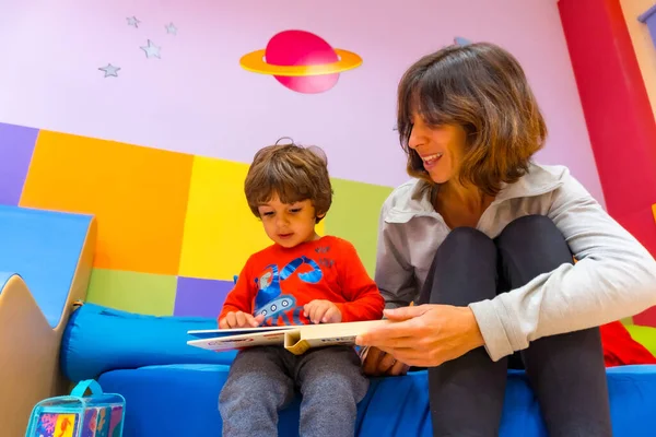 Kadın öğretmen bir çocukla oynuyor kitap okuyor ve bir anaokulunun içinde eğleniyor.