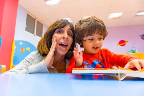 Çocuklu bir kadın öğretmen bir masada oturuyor ve bir anaokulunda masal kitabı okuyor.