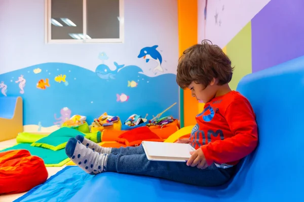 Mavi bir kanepede oturan bir çocuk bir anaokulunun içinde masal kitabı okuyor.